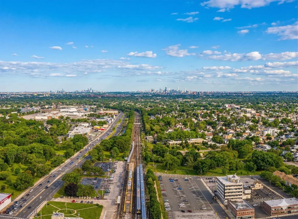 Aerial view of a suburban area with train tracks running through the center, surrounded by trees, houses, parking lots, and roads, with a distant skyline under a blue sky with scattered clouds.