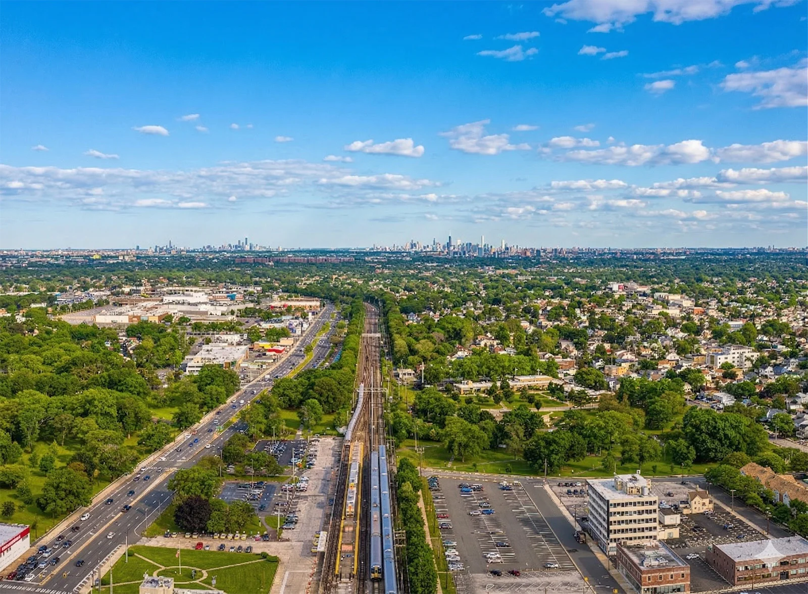 Aerial view of a suburban area with train tracks running through the center, surrounded by trees, houses, parking lots, and roads, with a distant skyline under a blue sky with scattered clouds.